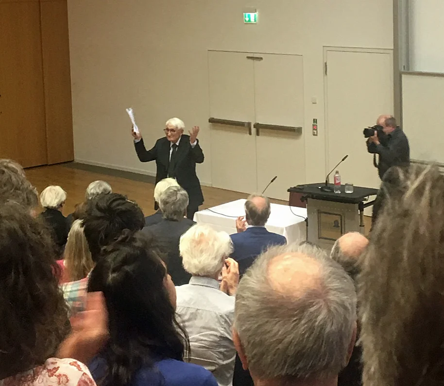 大学の大講義室の演壇で語るハーバーマス。段々の客席とマイク、投影スクリーンのある講演の風景。